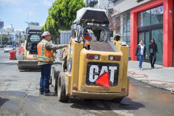 Worker operating yellow CAT skid steer loader on city street during utility work project.