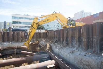Yellow excavator loading construction debris into a dump truck at an urban transit construction site.