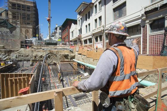 Construction worker in safety vest lowering rebar at Chinatown Station excavation site.