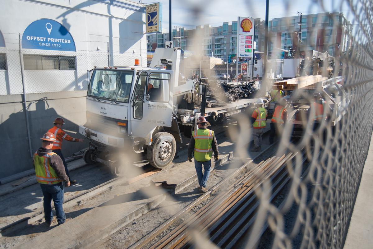 Construction workers in safety vests operate equipment near a truck at an urban transit construction site behind a chain-link fence.