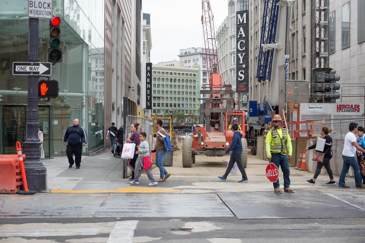 Construction work at Stockton and Geary intersection with heavy equipment, barriers, and flaggers managing pedestrian and vehicle traffic.