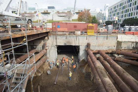 Construction workers organize rebar at Yerba Buena Moscone Station excavation site with city skyline in background.