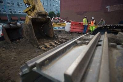 Excavator removing soil from trench with construction workers and equipment on site near buildings.