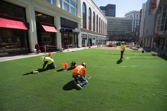 Workers in safety vests install artificial turf on a downtown San Francisco street surrounded by buildings.