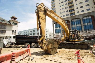 Yellow excavator at Stockton and Washington station site with construction barriers and nearby buildings.
