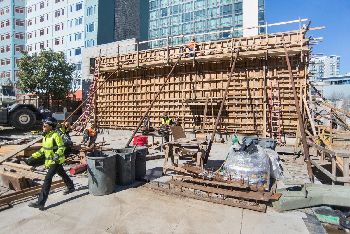 Construction workers assemble wooden scaffolding and framework at an urban transit station site.