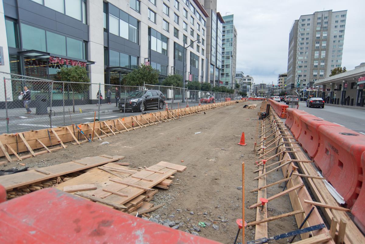 Construction site with excavation barriers, wooden forms, and red safety barriers along urban street with buildings.