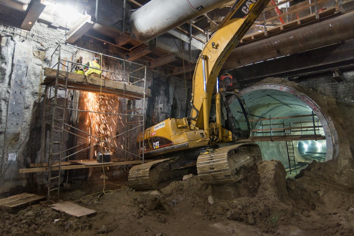 Yellow excavator operates inside underground tunnel construction site with worker welding steel framework.