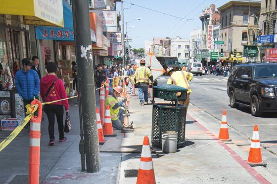 Workers in yellow vests check utilities on Stockton Street sidewalk with traffic cones and construction equipment visible.
