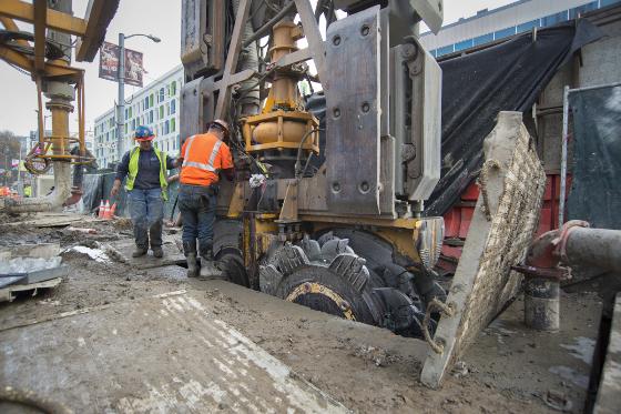 Two construction workers operate a hydromill machine on a city street during transit station construction.