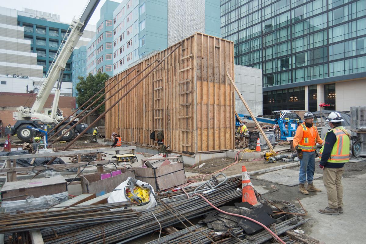 Construction workers assemble wooden formwork at an urban building site surrounded by high-rise buildings and equipment.