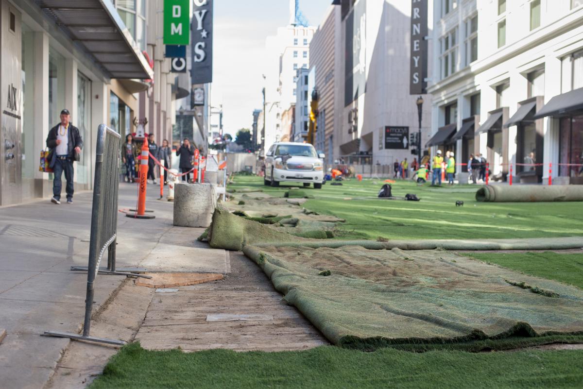 Green turf carpet installation along Stockton Street in downtown San Francisco with pedestrians and construction workers.