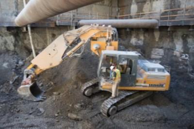 Yellow excavator lowering steel bracing into underground construction site near concrete structure.