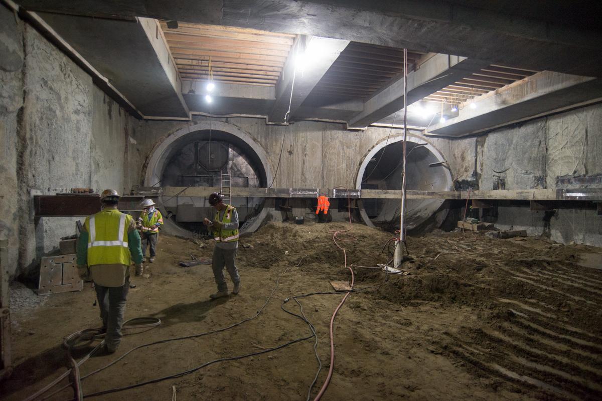 Workers in safety vests install large circular tunnel segments in underground construction site with steel bracing.