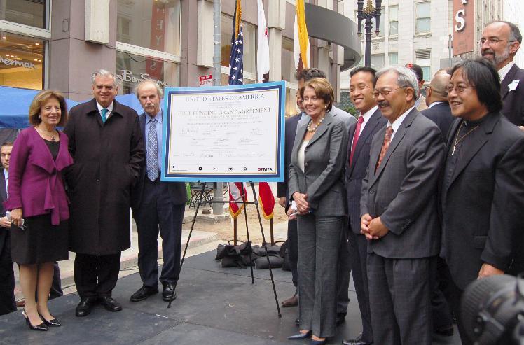 Federal, state and local officials display a framed grant award certificate at a downtown street ceremony with American flags in background.