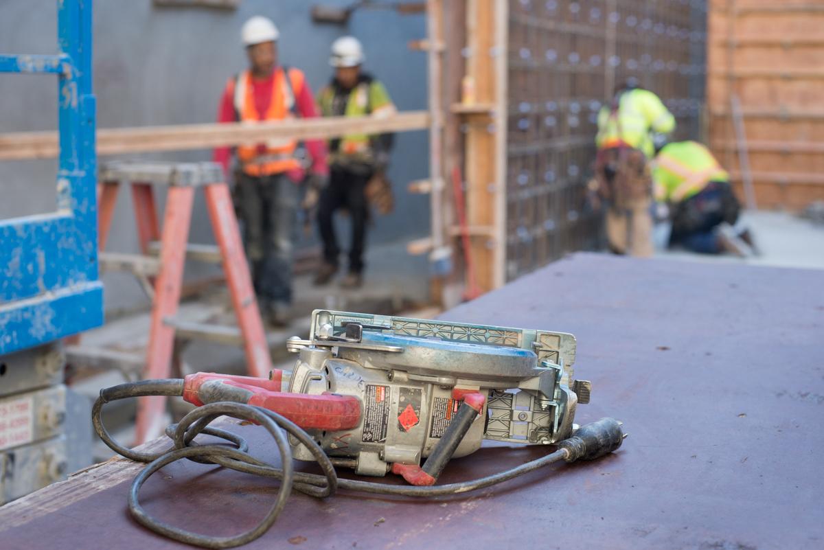 Construction workers building concrete forms at transit station headhouse, with power tools and safety equipment visible on site.