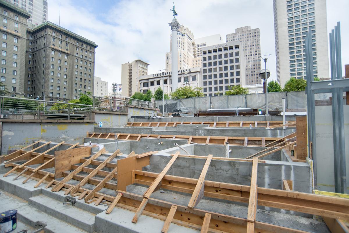 Construction site with wooden framing and concrete foundations for future plaza terraces near Union Square station, downtown skyline visible.
