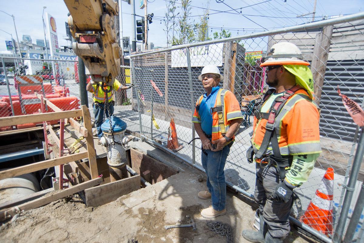 Construction workers in safety gear operate a crane to relocate a fire hydrant during Central Subway auxiliary water line work.