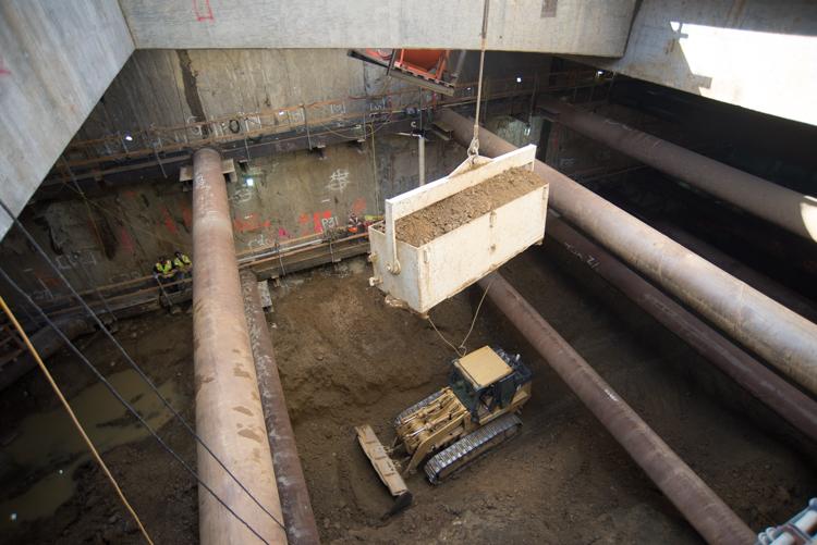 Excavation equipment removes soil from underground station construction site with large pipes and support structures visible.