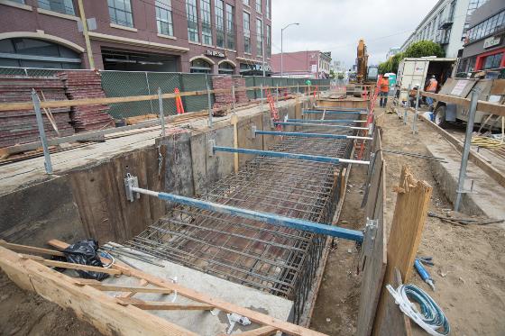 Construction site showing reinforced steel framework and wooden shoring in excavated pit on urban street with buildings in background.