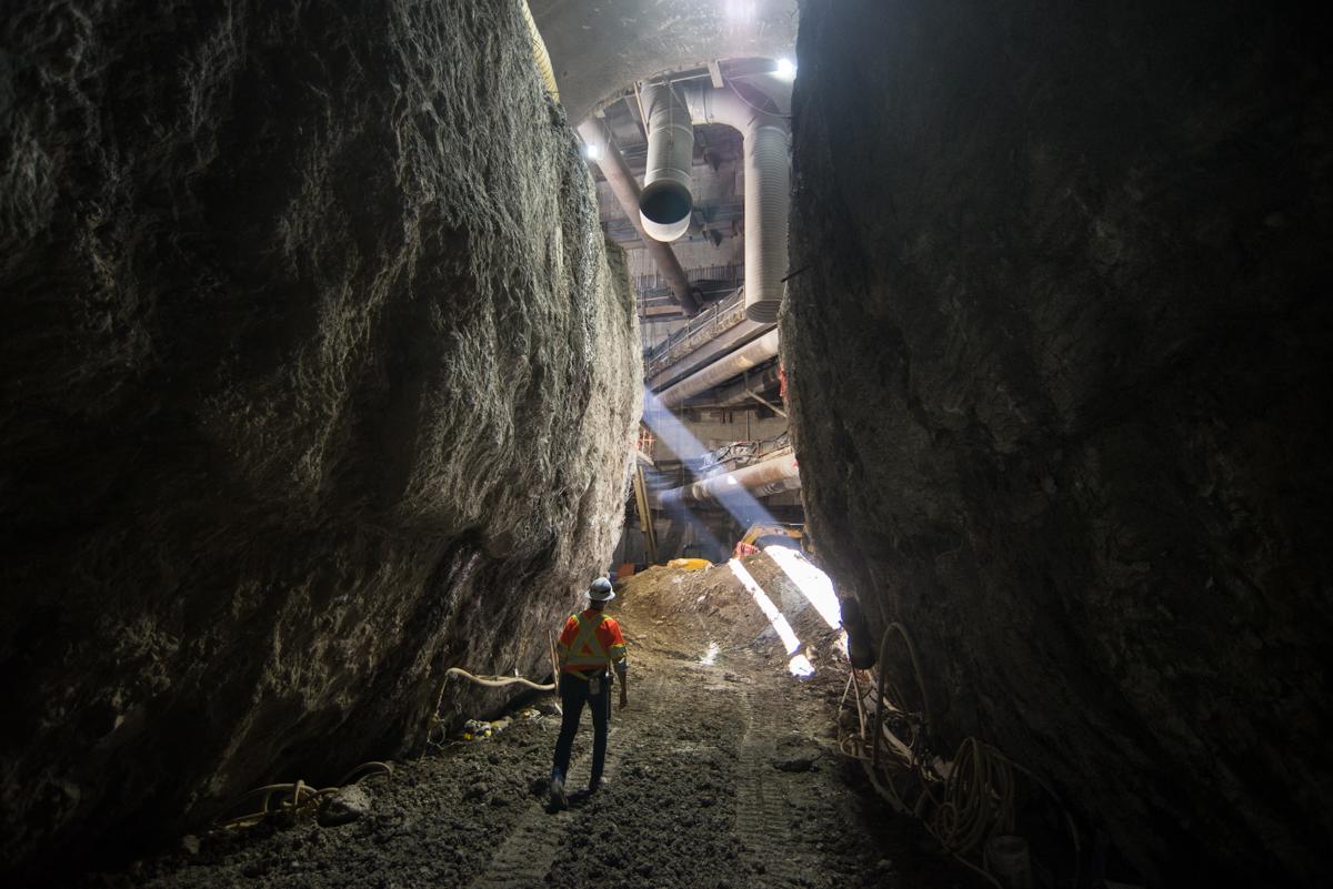 Worker in hard hat standing in underground tunnel with curved concrete walls and overhead pipes under construction lighting.
