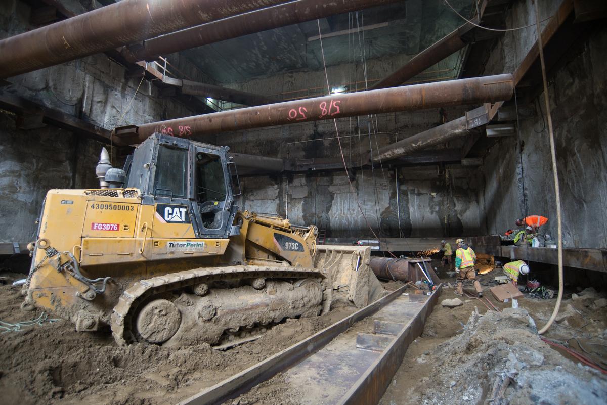 CAT bulldozer excavating platform level beneath large steel support beams in underground transit station construction.