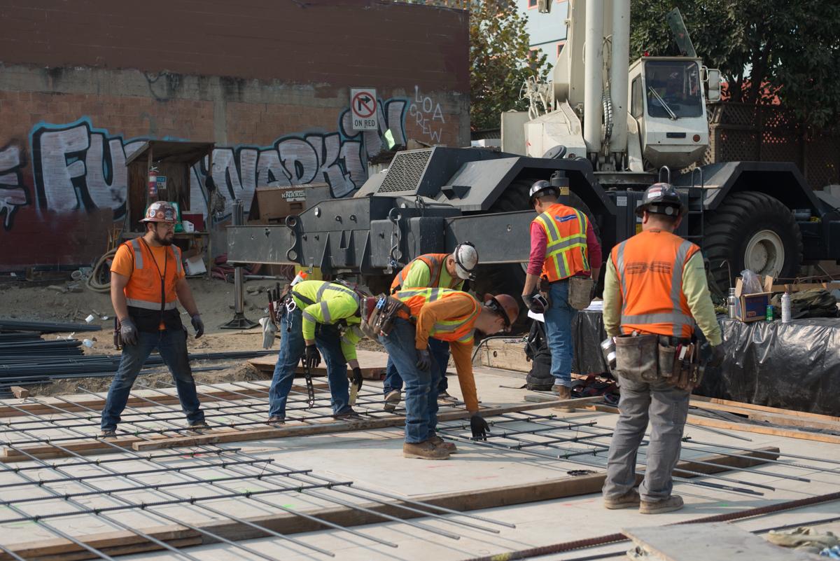 Construction crew assembling steel rebar reinforcement cages on a city street with heavy equipment nearby.