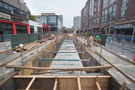Construction excavation pit along 4th Street with wooden framework and chain-link fencing, surrounded by urban buildings.