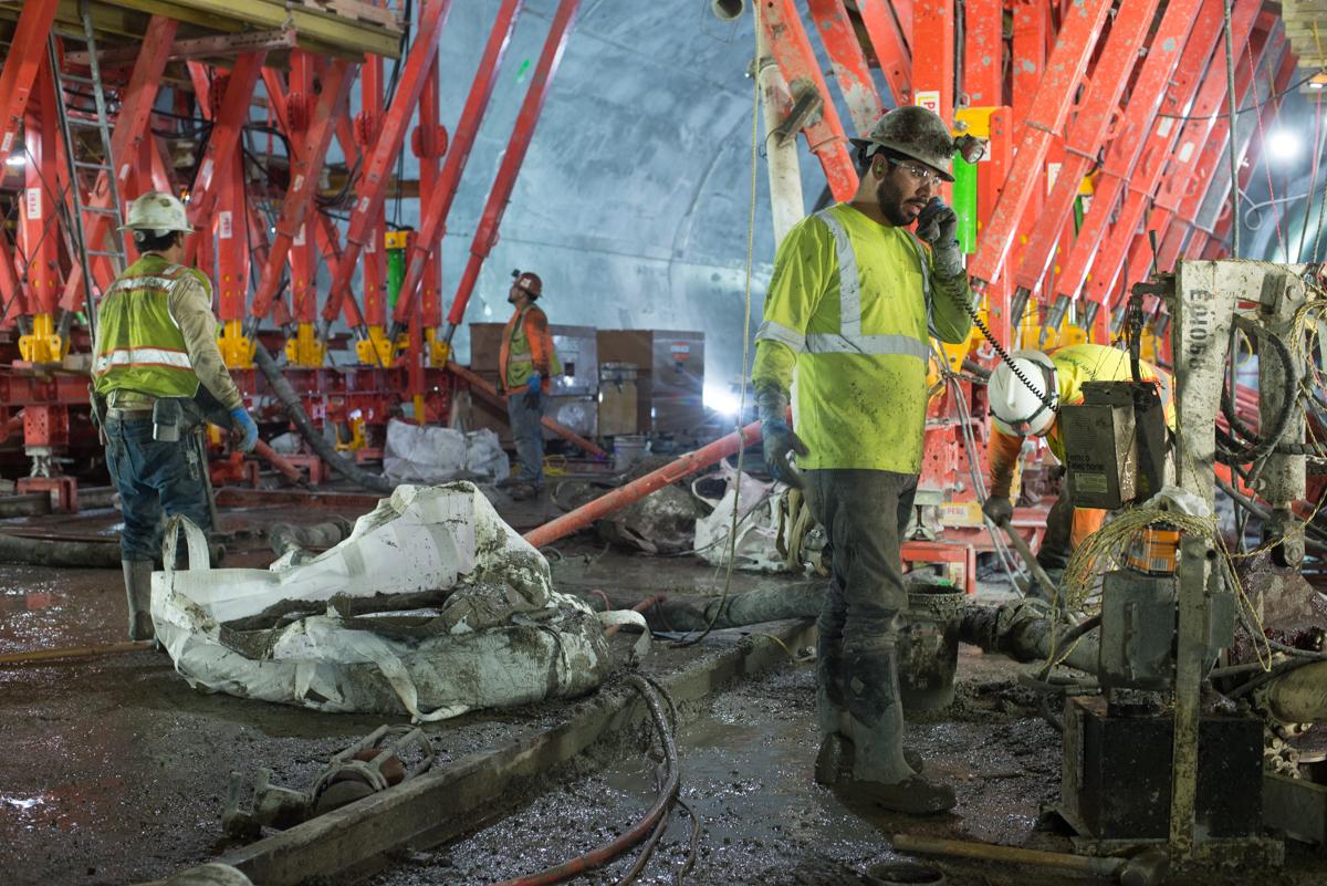 Workers in safety vests operate equipment inside a transit station under construction with red steel framework overhead.