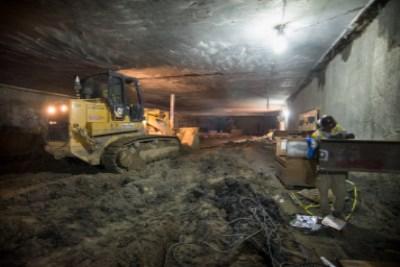 Bulldozer levels soil inside underground station box during nighttime construction work.