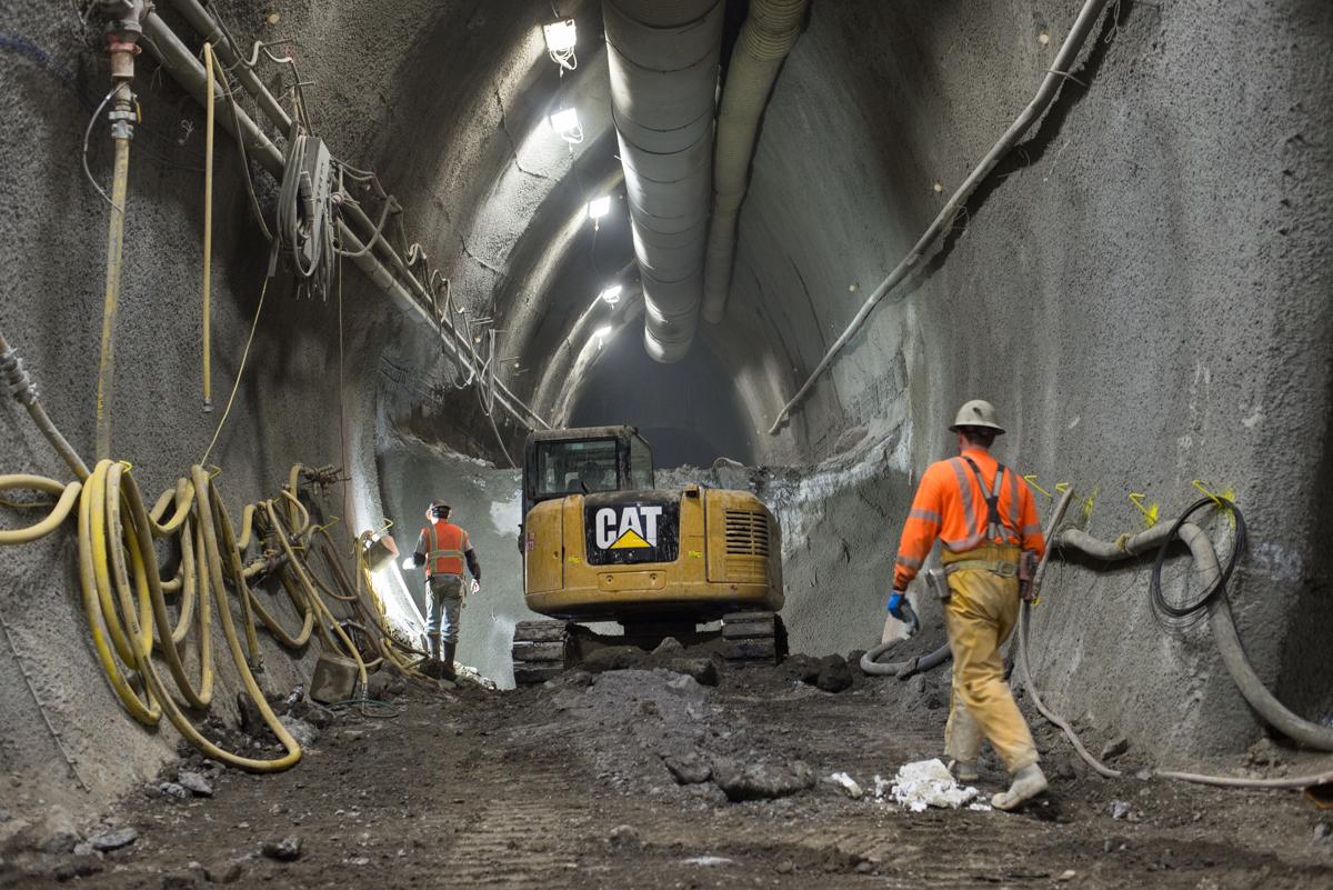 Workers in hard hats operate a CAT excavator in an underground tunnel during construction, with coiled cables and pipes visible along the concrete walls.
