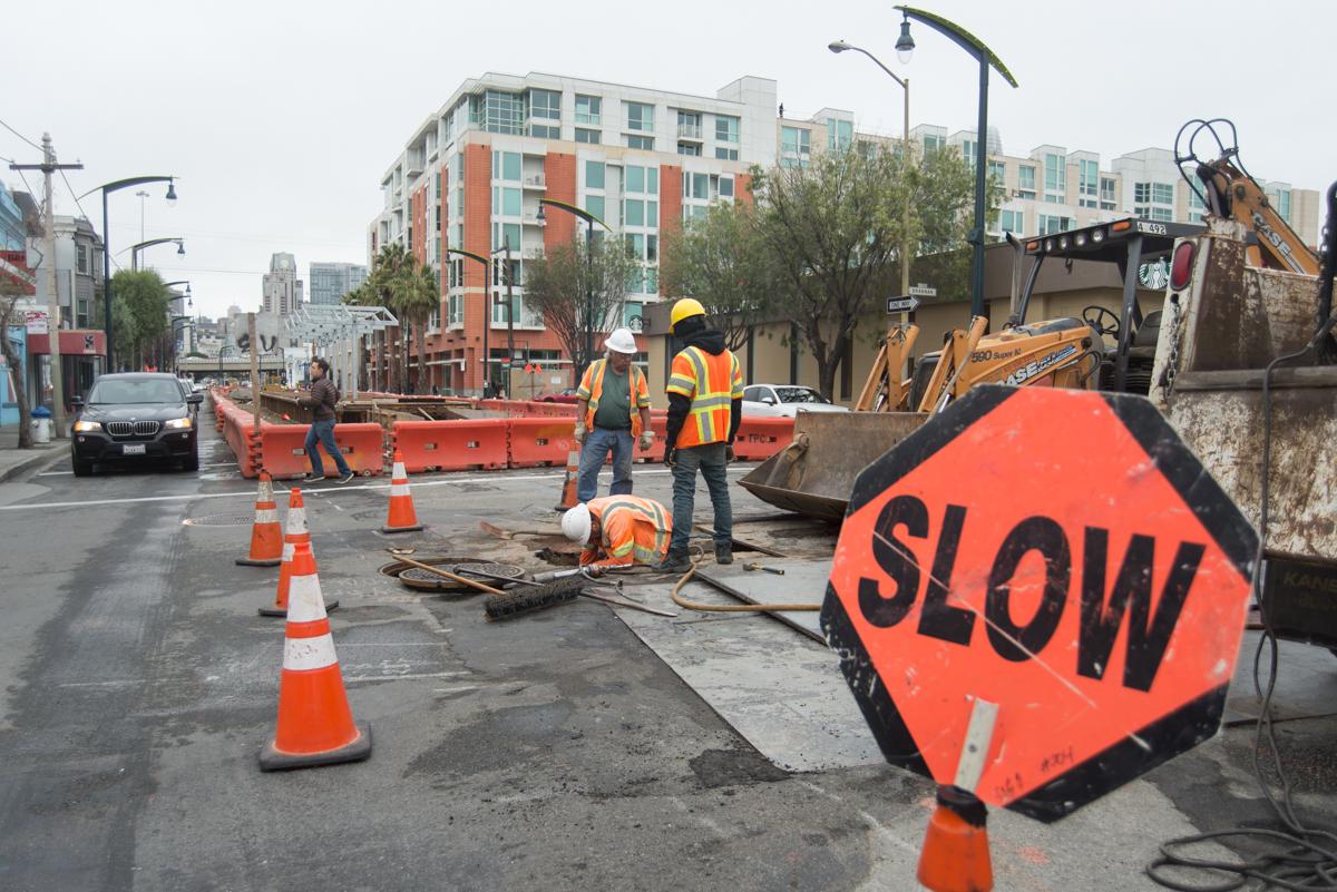Construction workers and equipment at 4th and Brannan intersection with traffic cones and slow sign during water line upgrade work.