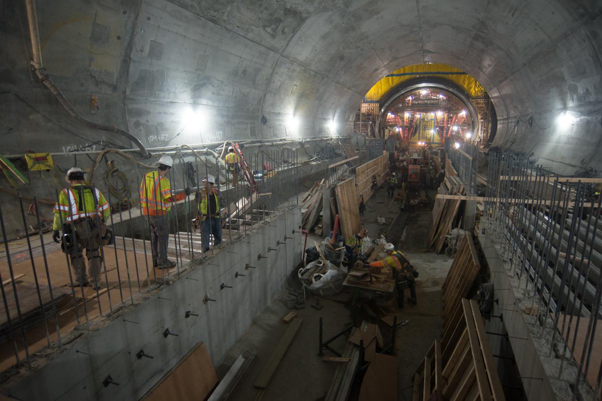 Construction workers in safety vests inside a large concrete tunnel during Central Subway station construction.