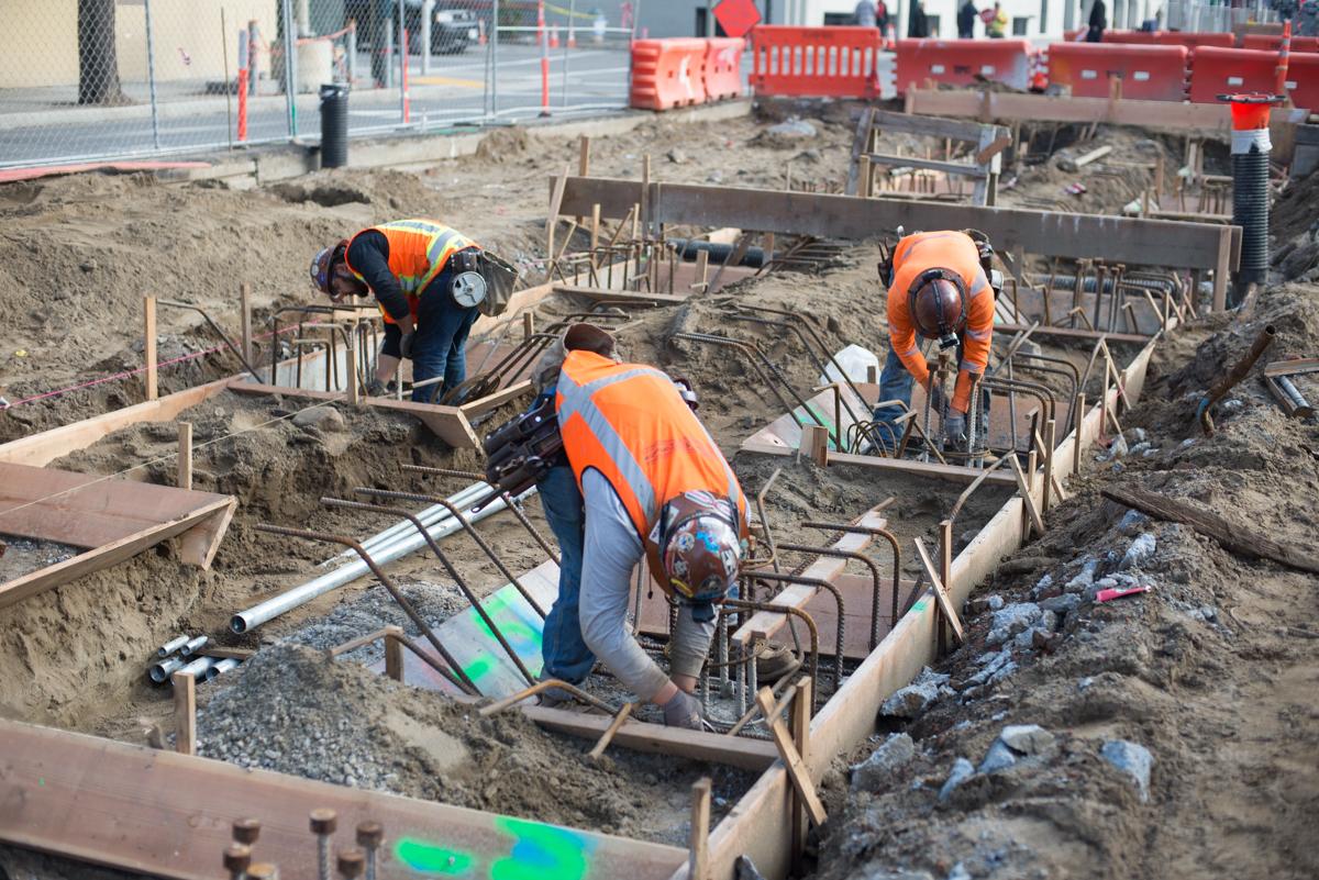 Construction workers in safety vests install rebar on foundation piles at a transit station construction site.