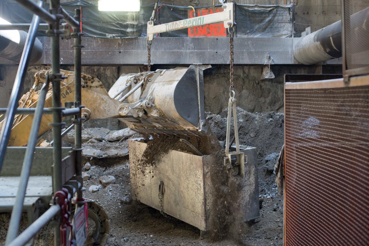 Concrete chute suspended by chains receives excavated soil from construction equipment at underground transit station work site.