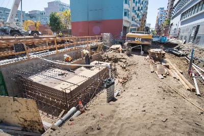 Construction site with excavation, utility pipes, and equipment on urban street with buildings in background.