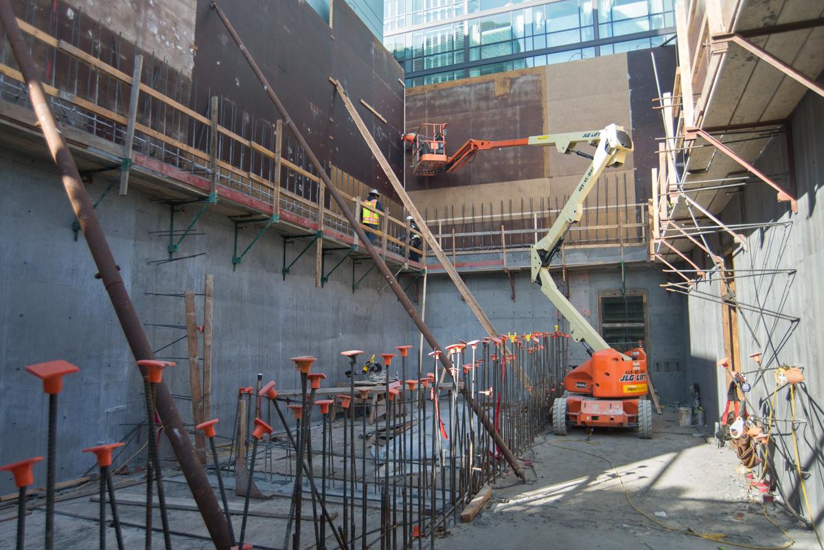 Construction site with concrete formwork, scaffolding, and orange aerial lift equipment inside an urban excavation pit.