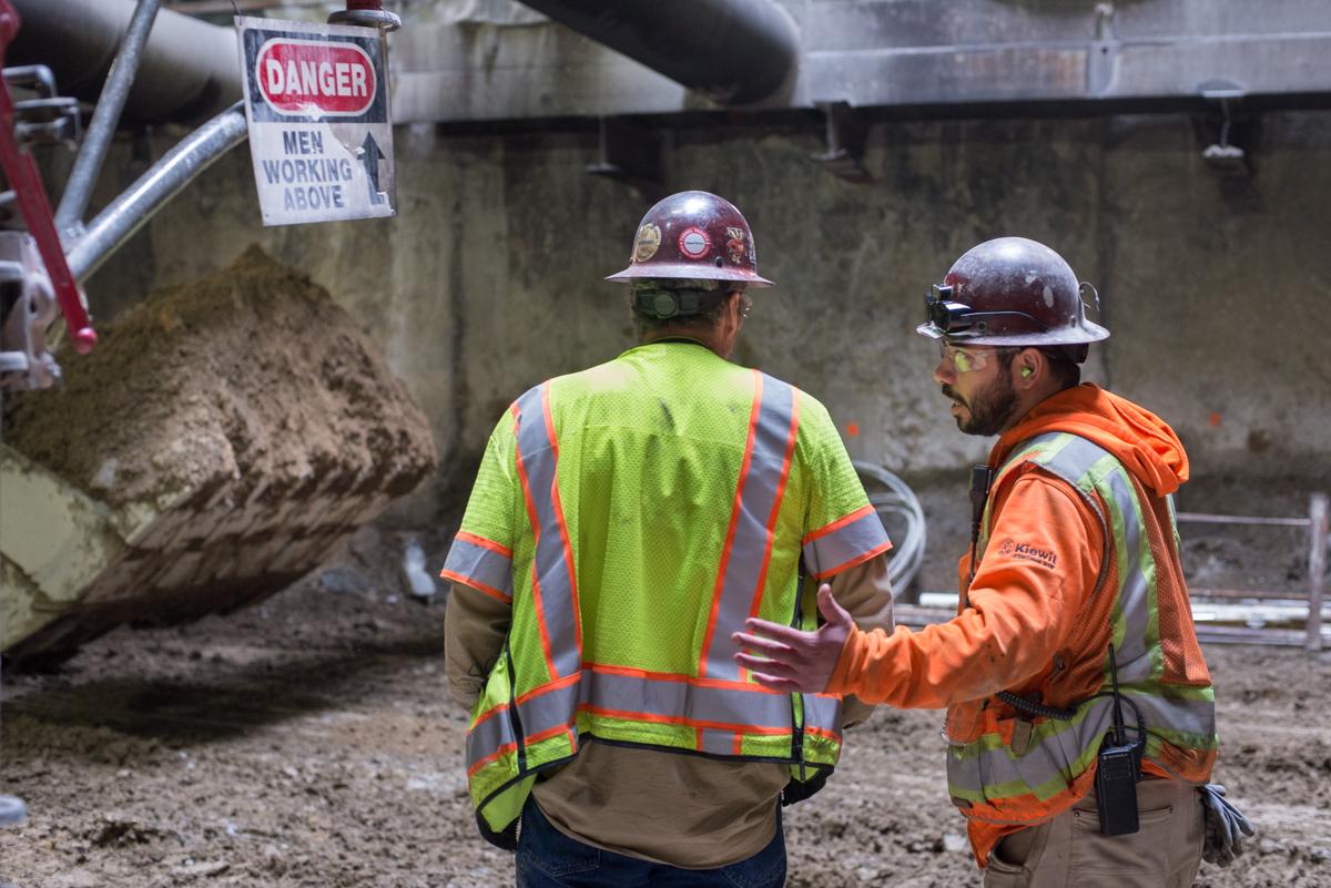 Two construction workers in safety vests and hard hats converse at an underground transit excavation site with a bulldozer nearby.