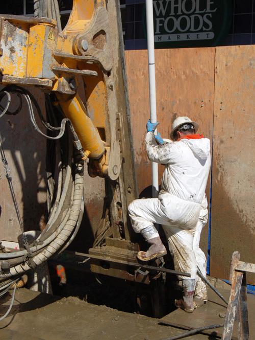 Worker in white protective suit operating concrete pump truck on 4th Street construction site near Whole Foods Market.