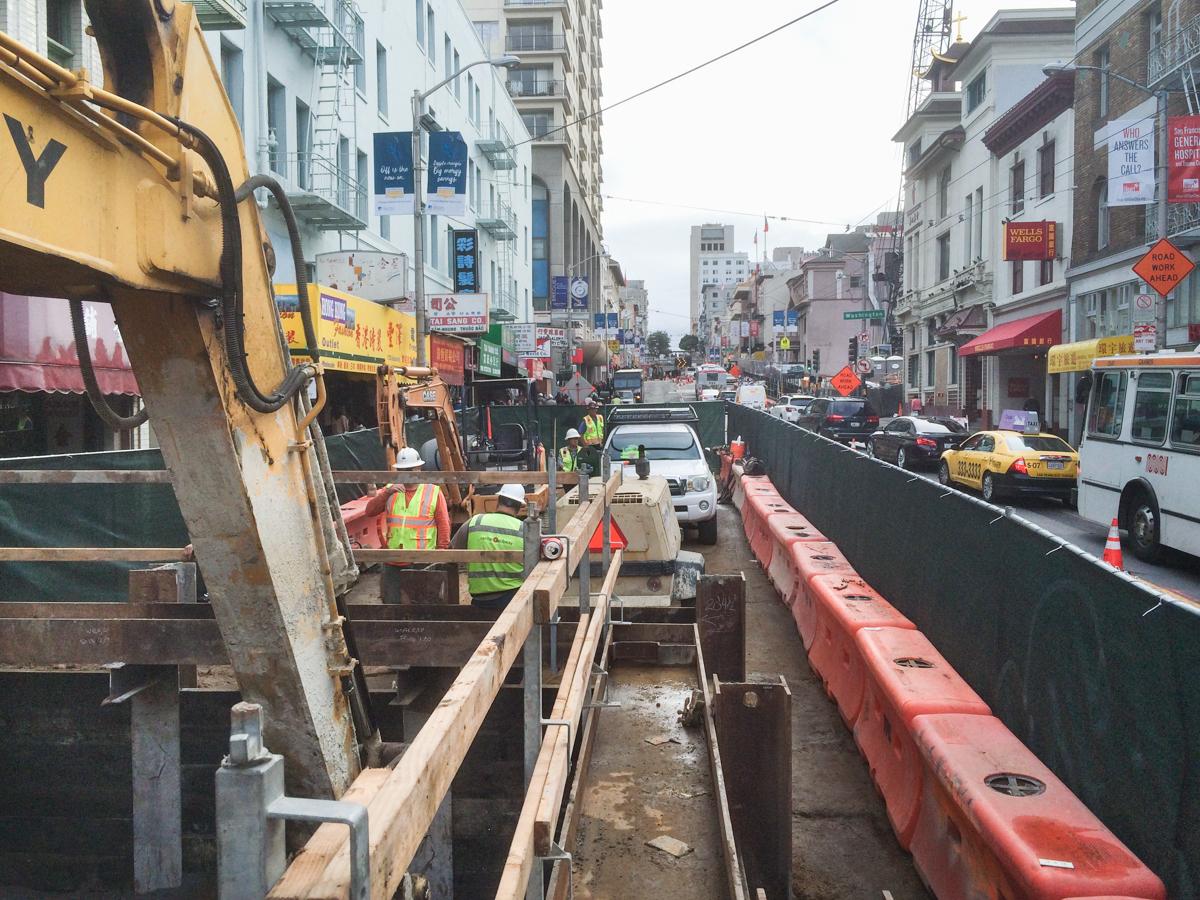 Construction excavation on Stockton Street with equipment, workers, and temporary barriers in urban area.