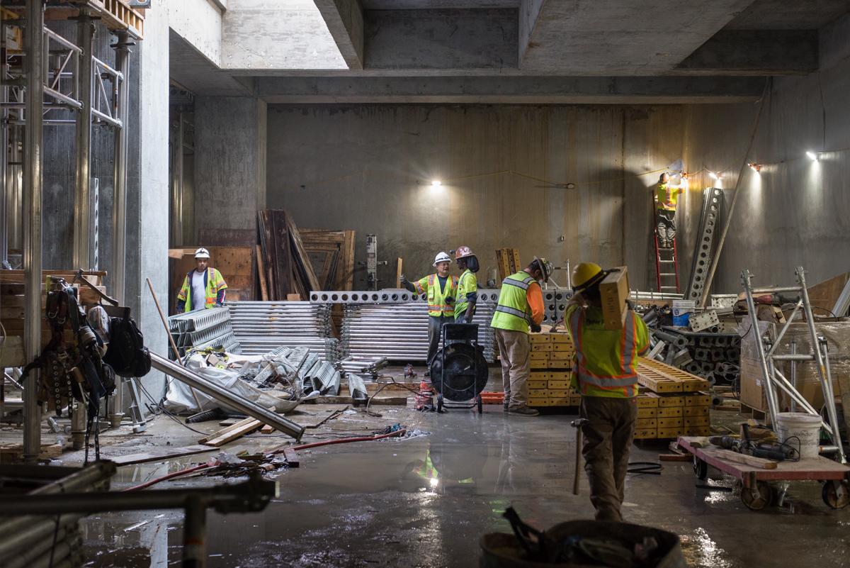 Construction workers in safety vests dismantle scaffolding and metal forms inside a concrete transit station under construction.
