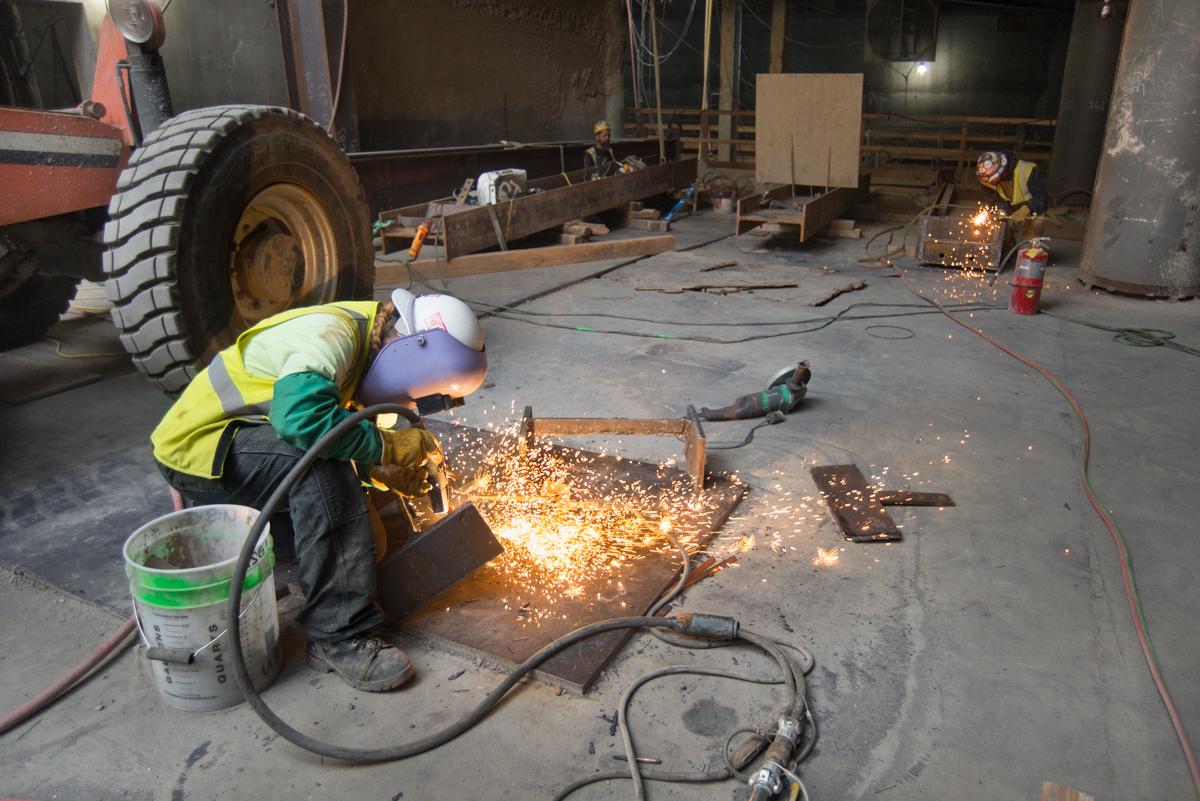 Worker in safety gear welding steel I-beams on concrete floor in underground transit station construction area.