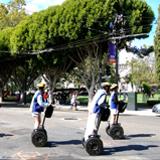 Two people riding electric scooters on a tree-lined street with parked cars and pedestrians in background.