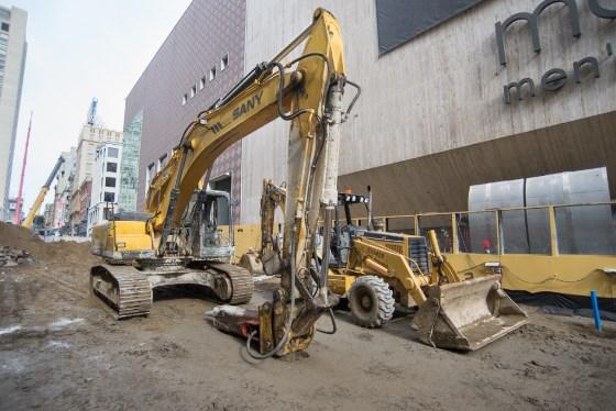 Yellow excavators and construction equipment on dirt lot surrounded by urban buildings.