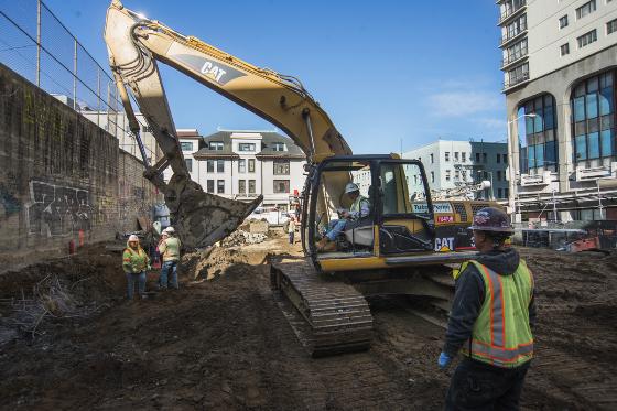 Construction crew excavates Chinatown Station site with CAT excavator and workers in safety vests.