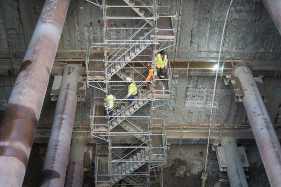 Workers in safety vests descend metal scaffolding and stairs in an underground transit station construction area.