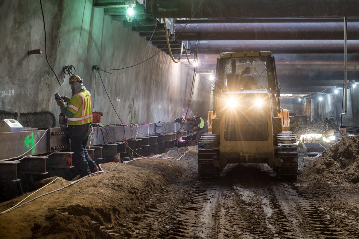 Workers in safety vests operate excavation equipment inside an underground transit station construction site.