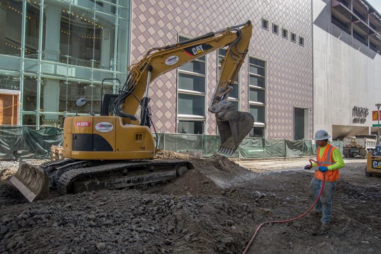 CAT excavator operates at Stockton Street construction site while worker sprays water to control dust.