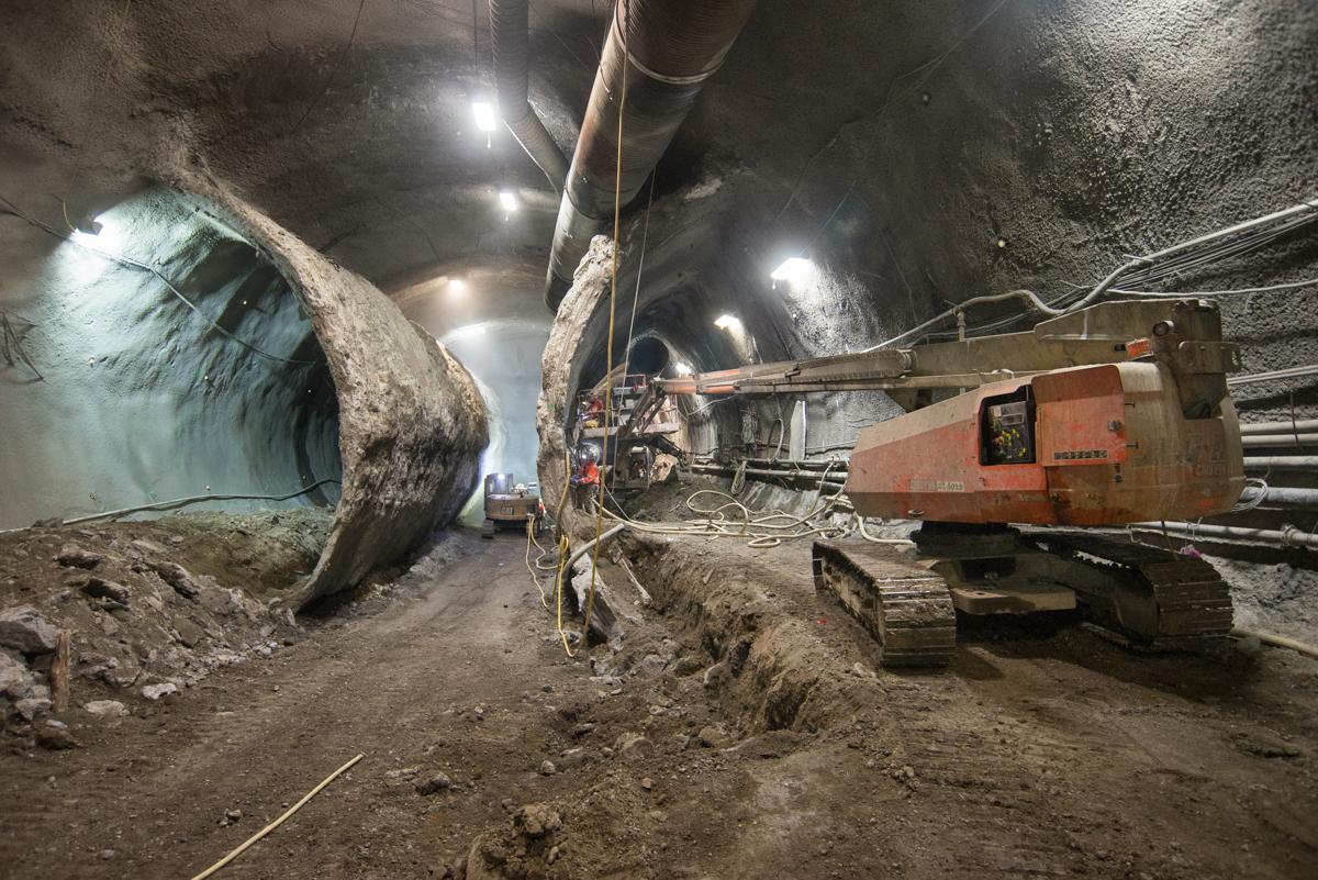 Excavation equipment operates inside an underground tunnel with concrete walls and overhead lighting during Central Subway construction.