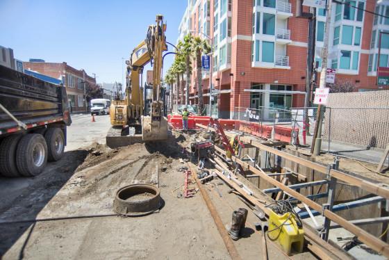 Construction excavator installing underground sewer infrastructure on urban street with buildings nearby.